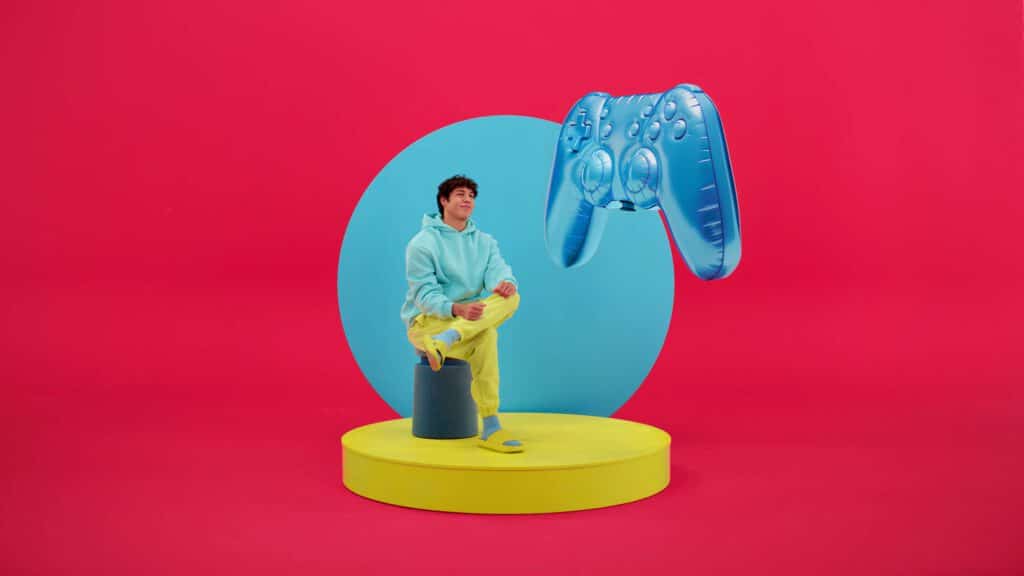 A person in colorful clothes sits cross-legged on a yellow platform with a large blue inflatable game controller floating beside them, set against a bright red and blue background—an eye-catching scene worthy of AI Art. AI Image made by Christian Fleischer / The Visiblemaker AI Advertising Campaign