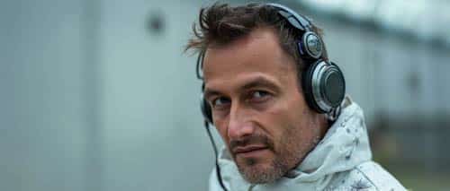 A man wearing a white patterned jacket and large headphones looks directly at the camera with a serious expression; the blurred gray background gives the scene an AI Cinema feel. AI Image made by Christian Fleischer / The Visiblemaker