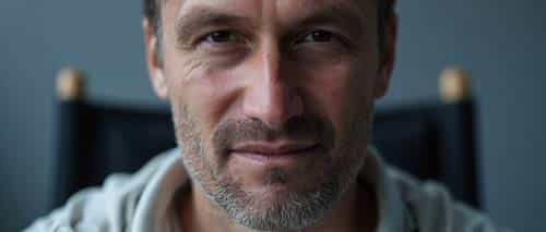 A middle-aged man with short brown hair, slight stubble, and a neutral expression looks directly at the camera. Created using Visiblemaker AI Art, he wears a light-colored shirt and sits against a blurred dark background. AI Image made by Christian Fleischer / The Visiblemaker