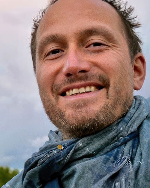 A man with short brown hair and a beard is smiling at the camera. He is wearing a blue scarf and standing outdoors, with a cloudy sky and some greenery in the background—an image created using AI Art by Visiblemaker. AI Image made by Christian Fleischer / The Visiblemaker