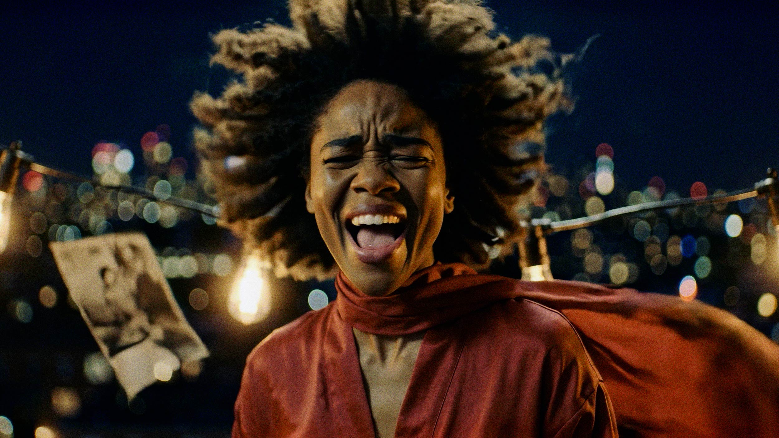 A person with expressive facial features and wild hair stands outdoors at night, wearing a red outfit with a flowing scarf. Blurred city lights and hanging bulbs glow in the background, evoking the vibe of a full ai musicvideo. AI Image made by Christian Fleischer / The Visiblemaker